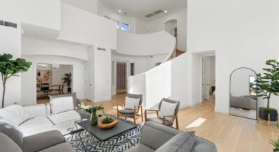 A modern living room in Gainey Ranch with white walls and light wooden flooring features a gray sectional sofa, armchairs, a black coffee table, a large mirror, and plants. The room has a high ceiling and a staircase.