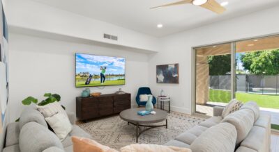 A modern living room in Gainey Ranch features a large couch, round coffee table, wall-mounted TV, ceiling fan, and glass doors leading to a serene backyard with a pool.