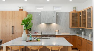 A modern kitchen in Gainey Ranch, featuring light wooden cabinets, a marble countertop and backsplash, bar stools, and a kitchen island adorned with a large plant, cutting board, glass bottles, and limes.