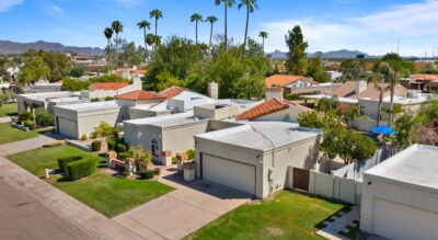 From an aerial view, the suburban charm of 8725 E San Marino Dr in McCormick Ranch unfolds with single-story houses and flat roofs dotted by palm trees. The picturesque mountain range stands majestically in the background against a clear blue sky.