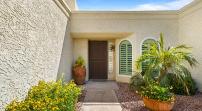 The entrance of 8725 E San Marino Dr McCormick Ranch features a stucco façade with a dark door, gracefully framed by potted plants and elegant arched windows. The surrounding gravel and vibrant flowering shrubs complete the picturesque scene.