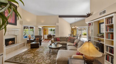 Nestled in the heart of McCormick Ranch, the spacious living room at 8725 E San Marino Dr features a beige sectional sofa, inviting fireplace, sleek coffee table, stylish rug, and elegant bookshelves. Sunlight pours through large windows, with an open dining area gracefully visible in the background.